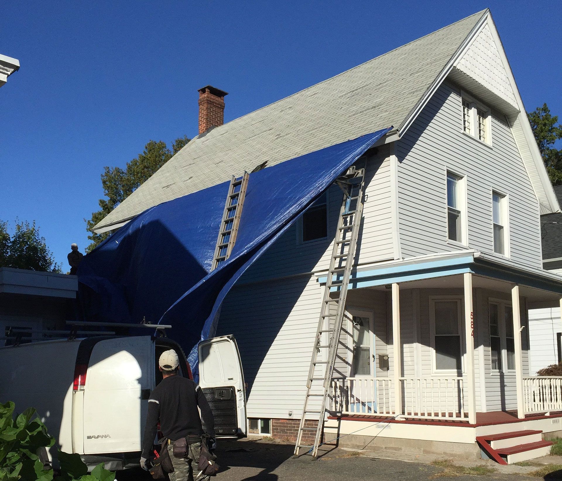 A worker uses a ladder to secure a large blue tarp over the roof of a two-story white house with a front porch.