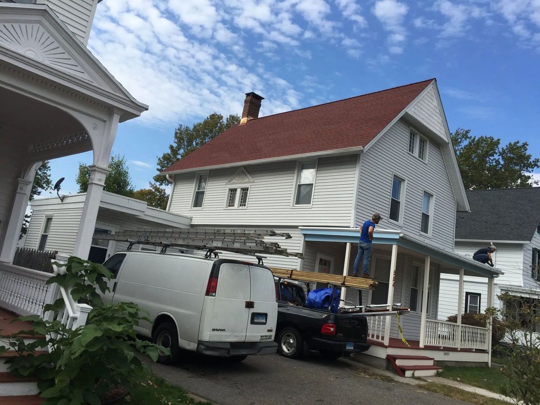 A construction crew works on the roof of a white house with a red roof, with a van and truck parked in the driveway.
