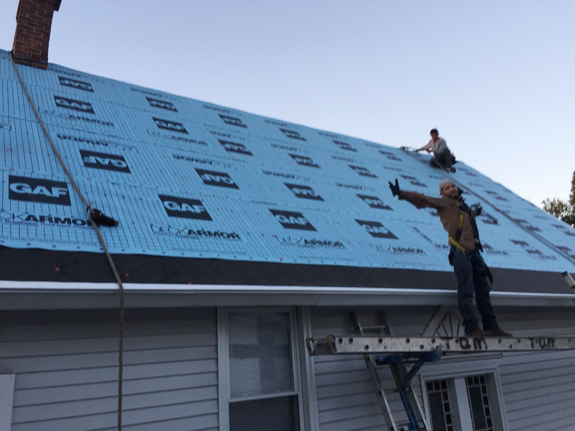 Two workers wearing safety harnesses install light blue GAF underlayment on a steep residential roof.