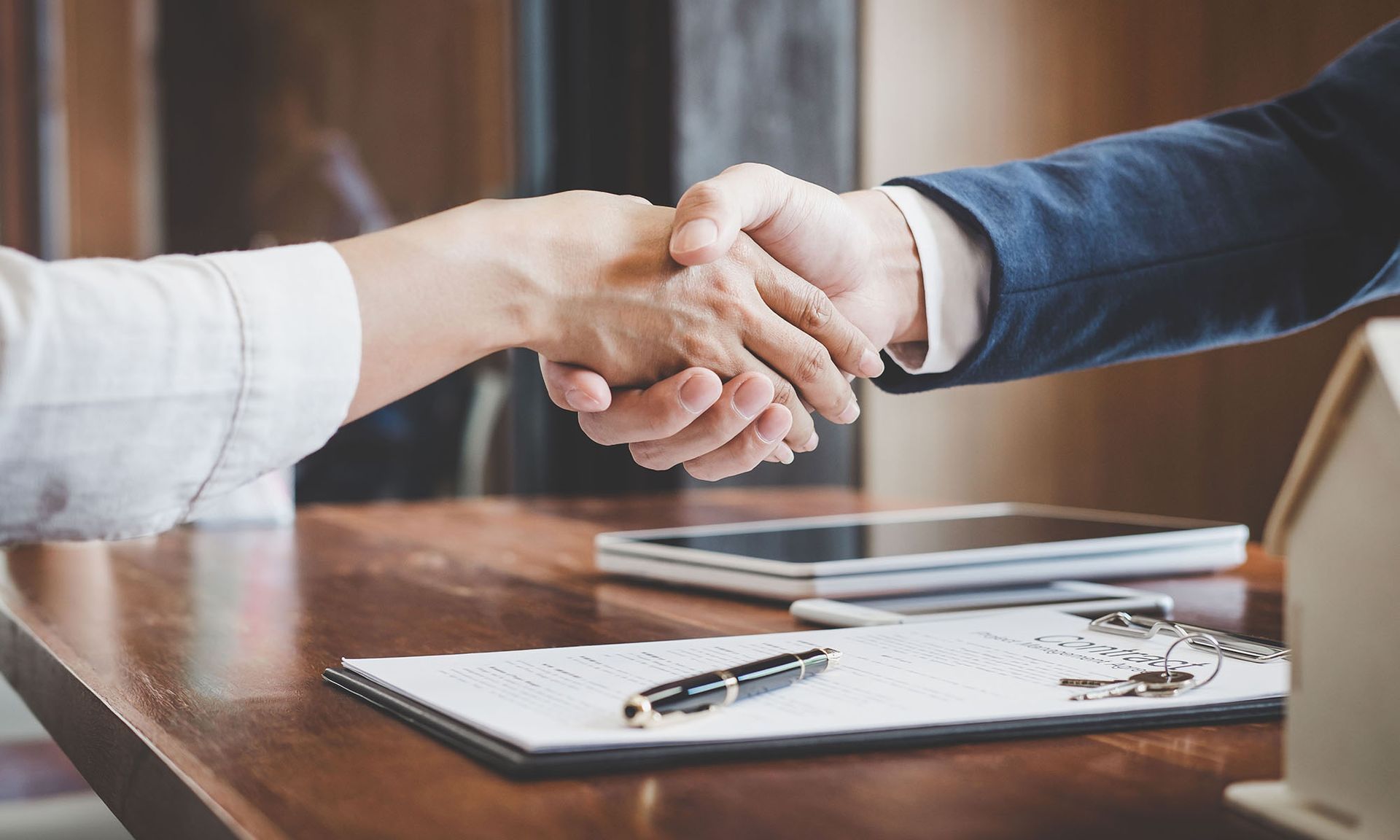 Two people shaking hands over a contract on a table; a deal is made.