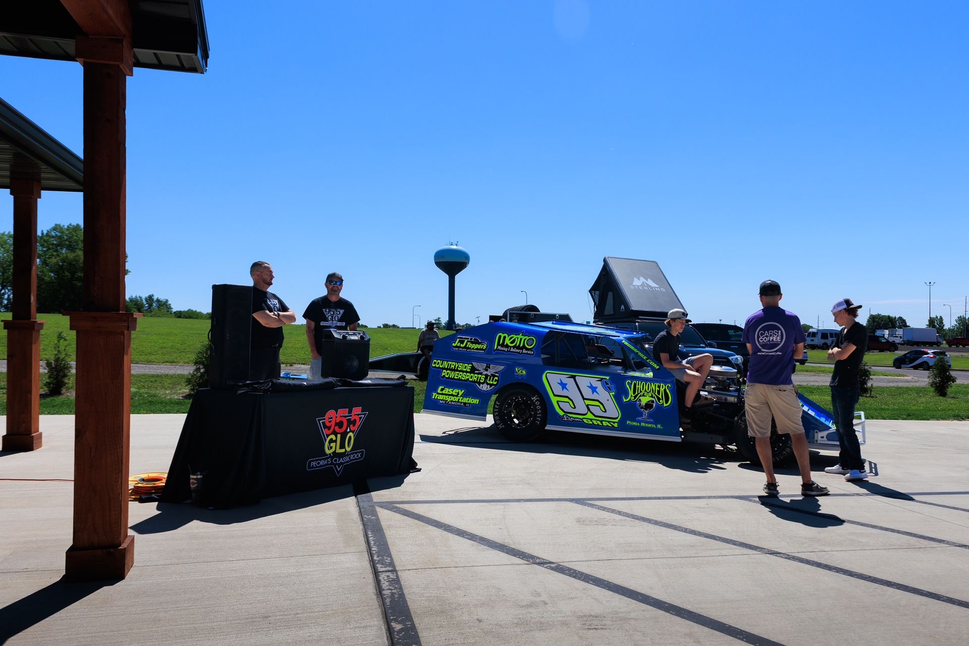 Race car with number 82 parked outdoors with several people standing nearby under a bright blue sky.