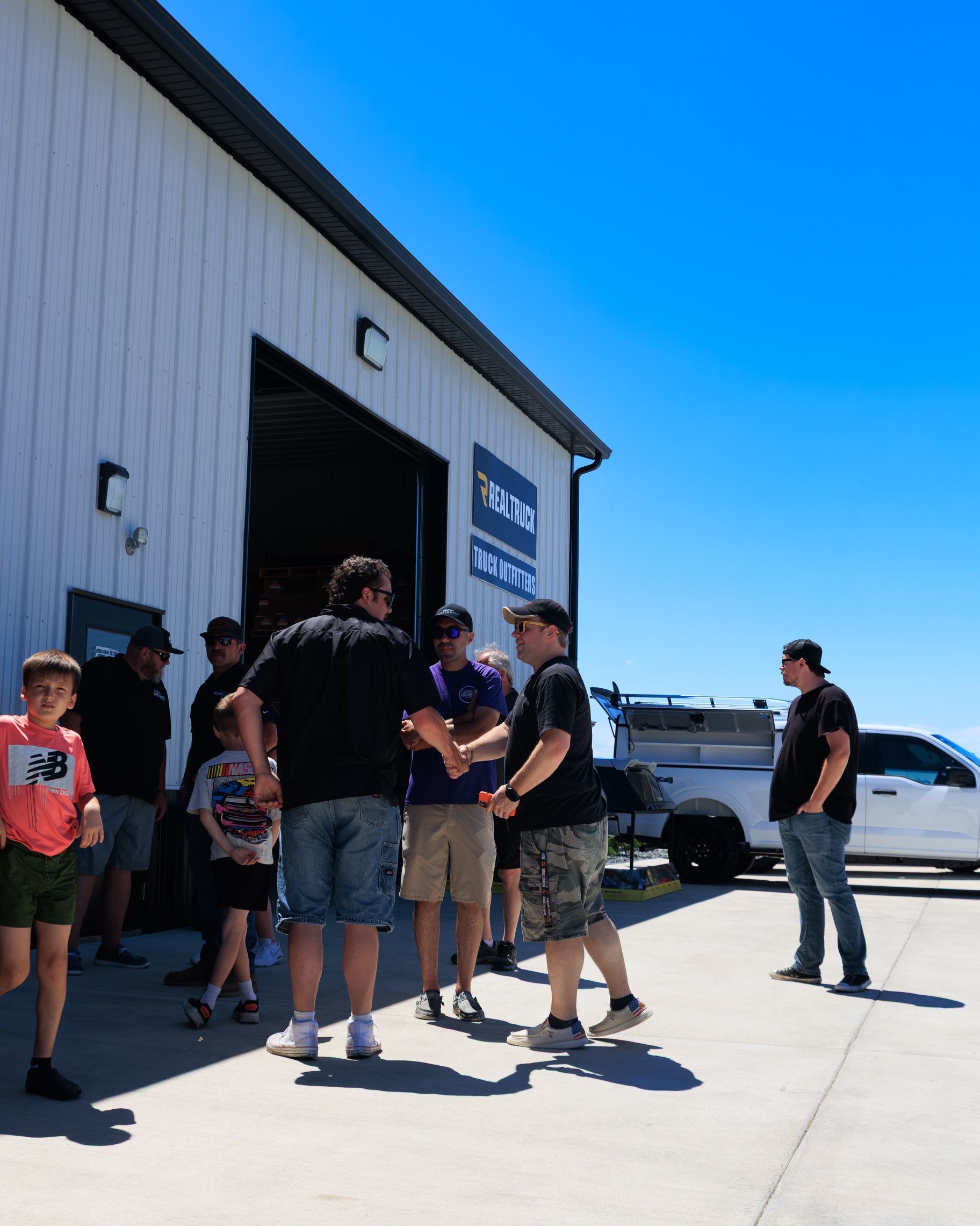 People gathered outside a warehouse, chatting near parked vehicles under a bright blue sky.