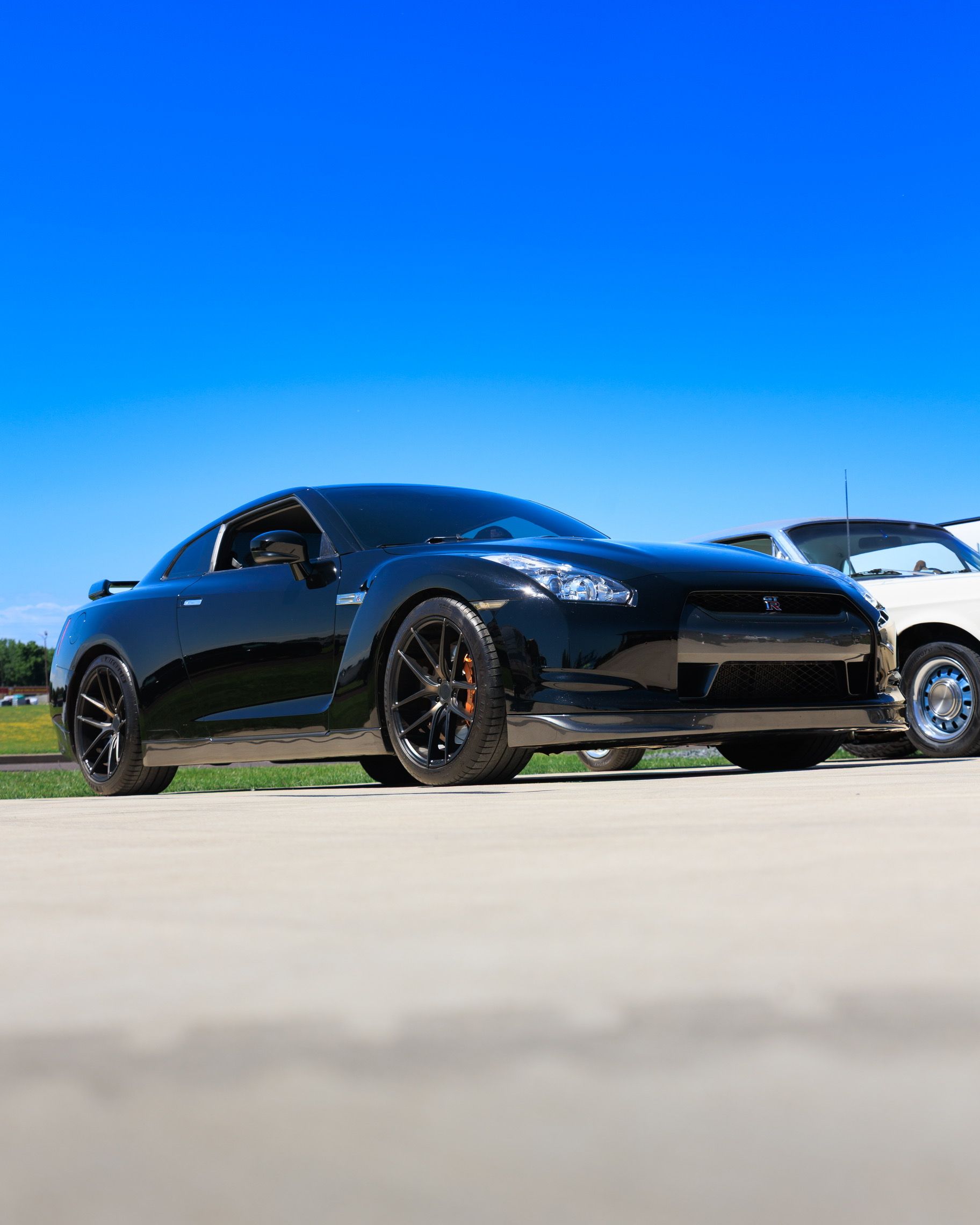 Black sports car parked outdoors under a bright blue sky, with a white car behind it.