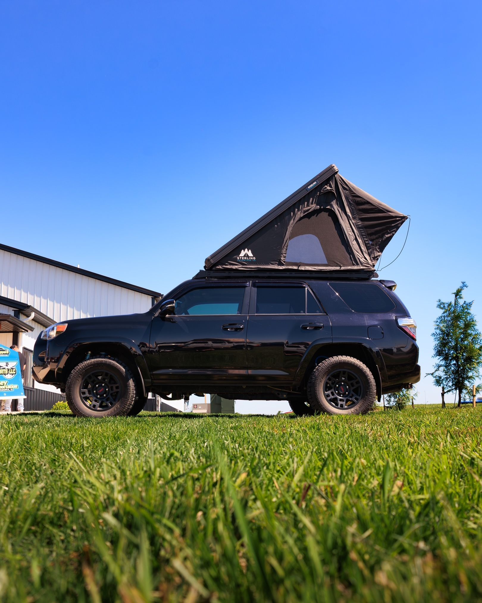 Black SUV with rooftop tent parked on grass under a blue sky.