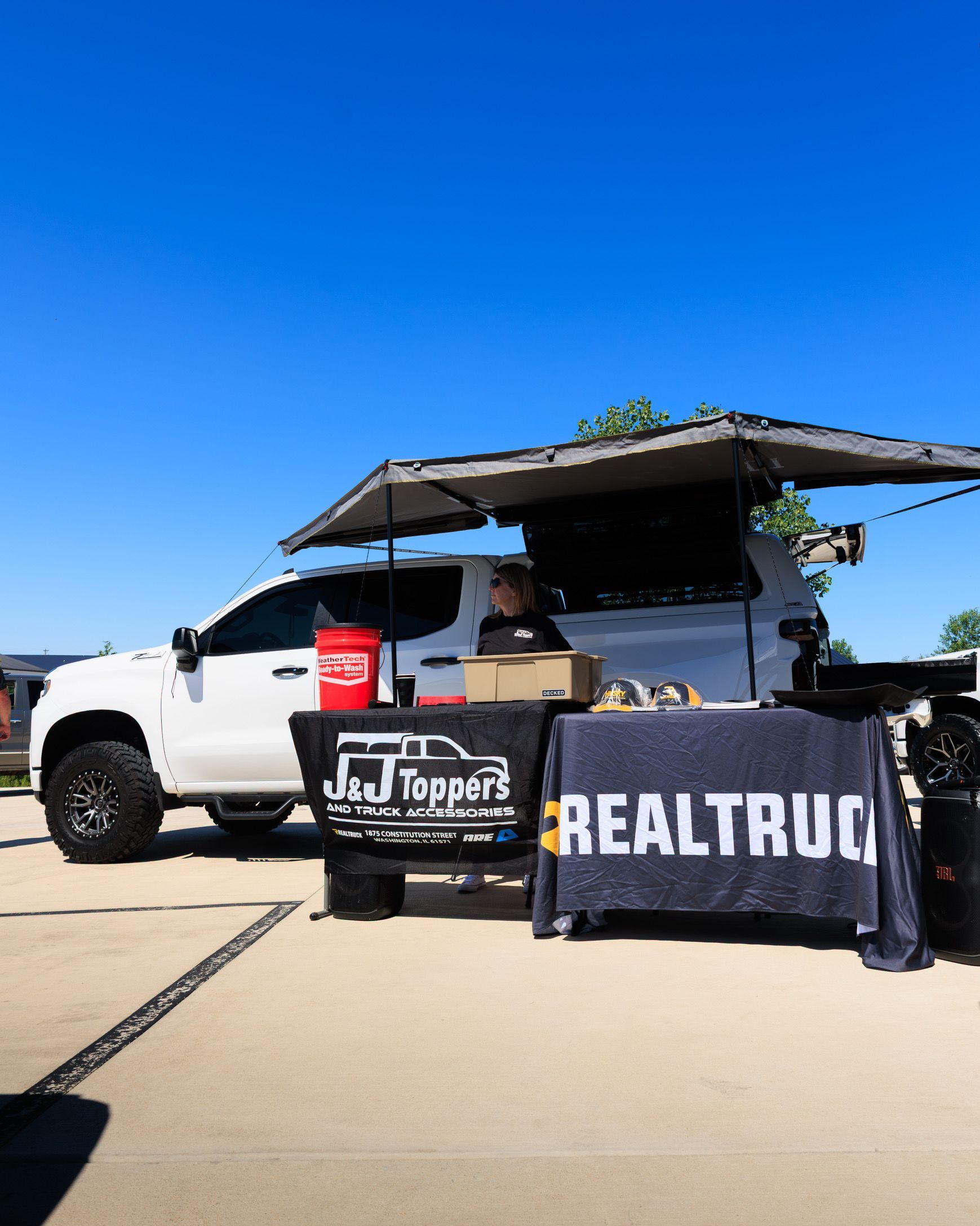White pickup truck with canopy, black REALTRUCK banner, and Topper display at an outdoor event under a blue sky