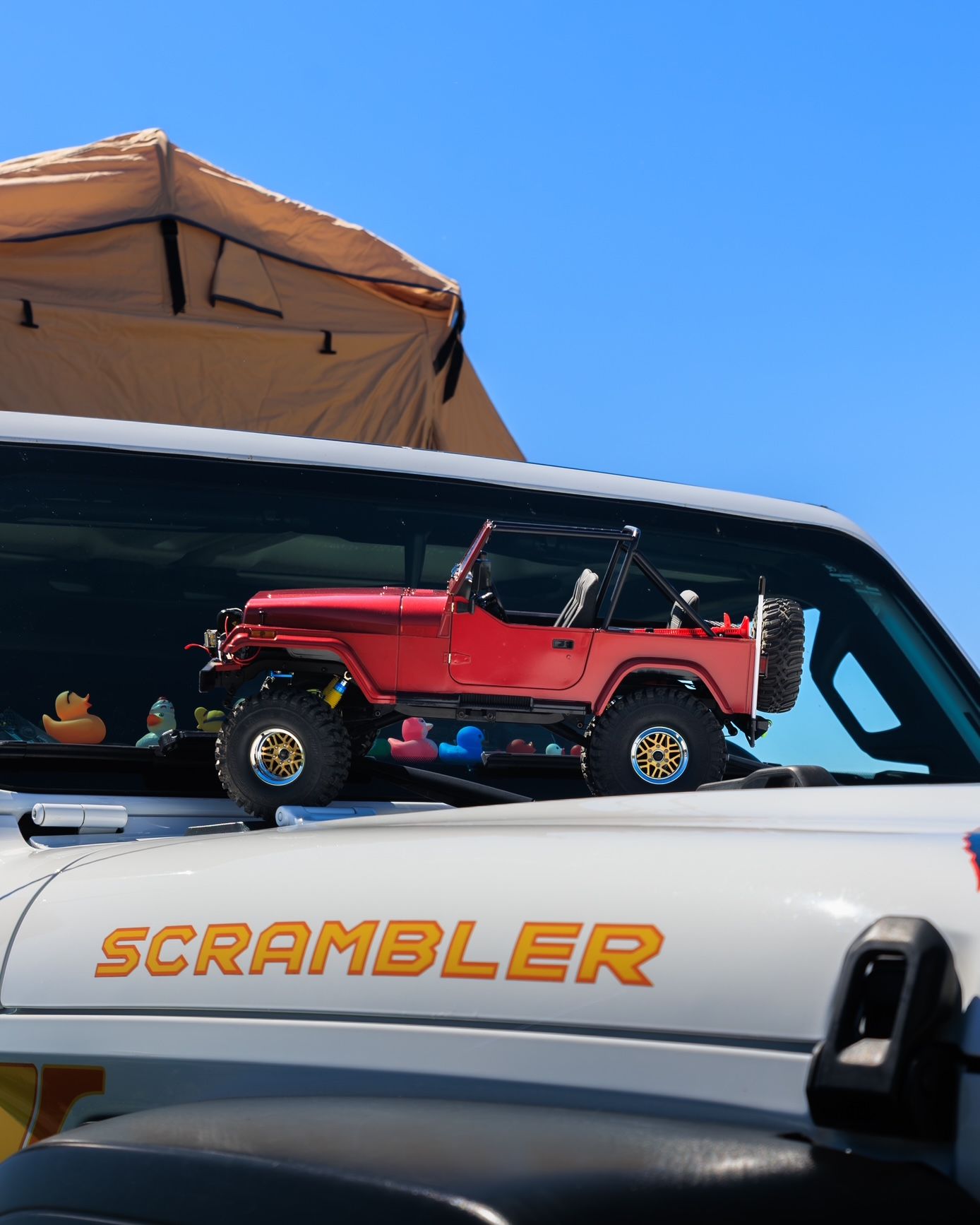 Red toy Jeep on a white vehicle hood with “SCRAMBLER” text, under a tan rooftop tent against blue sky