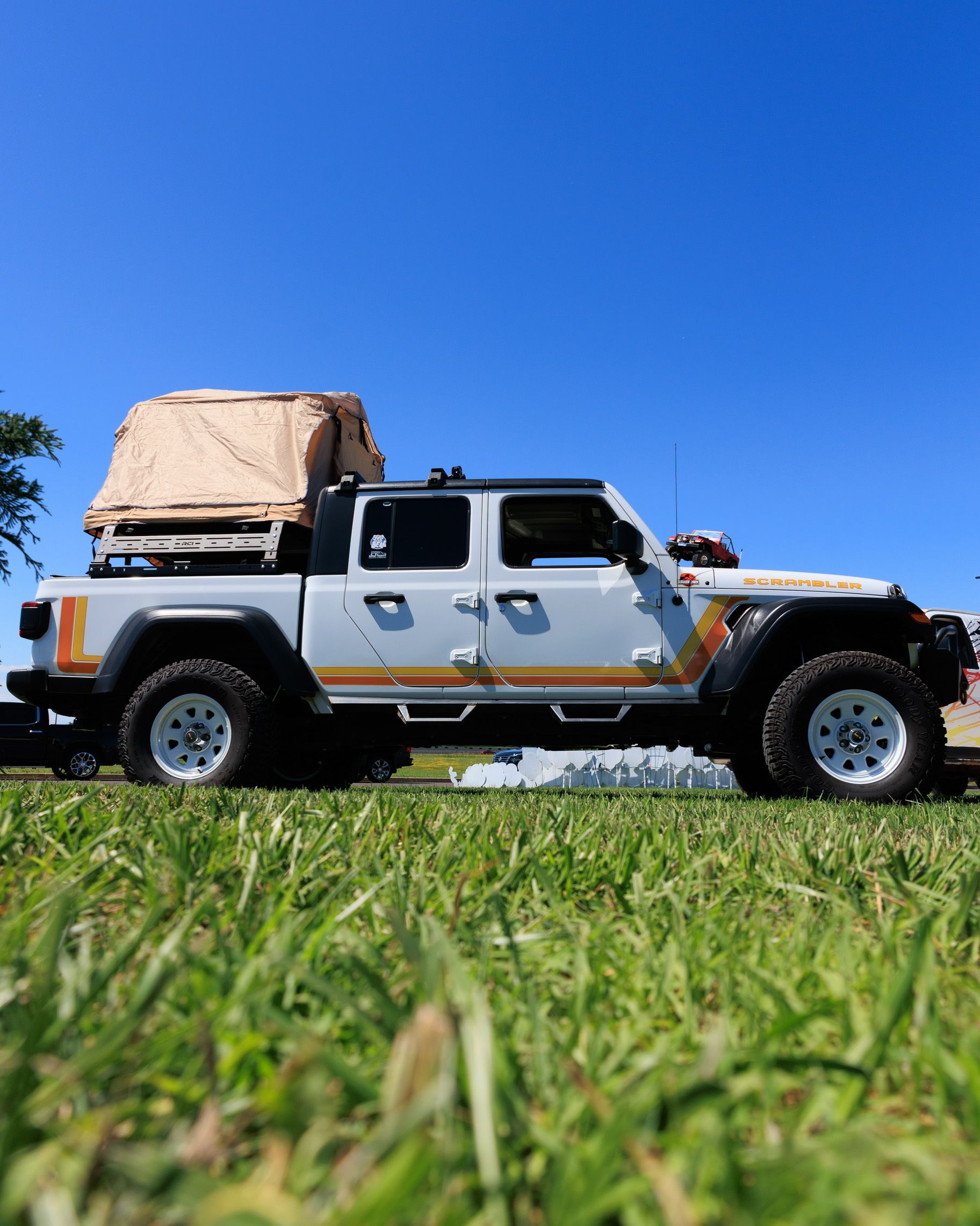 White off-road Jeep with rooftop gear parked on grass under a clear blue sky.