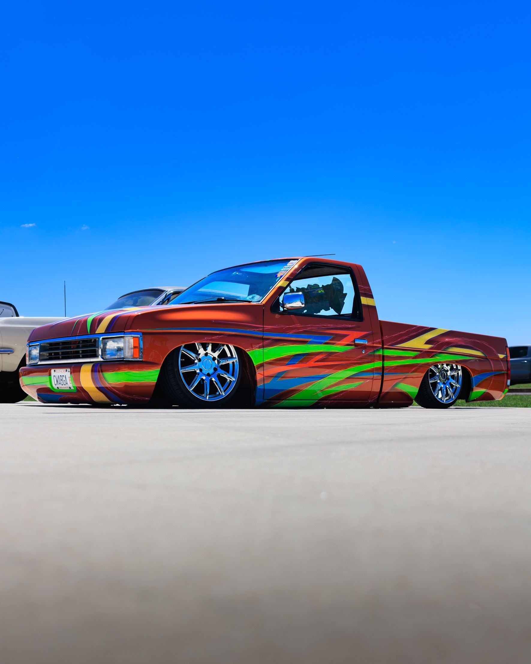 Custom lowrider pickup truck with vivid orange-green paint, parked on a road under a bright blue sky.