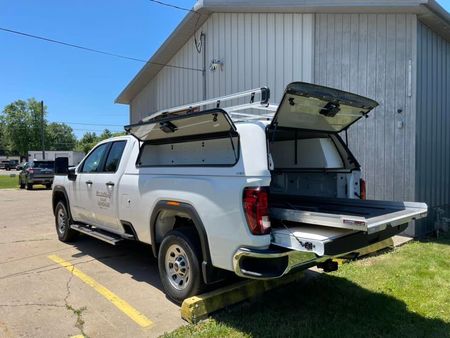 White truck with open canopy and cargo bed, parked on pavement next to grass, in front of a building.