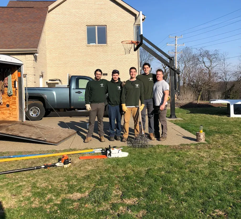 Four people standing by a house driveway with a pickup truck, lawn equipment, and a basketball hoop