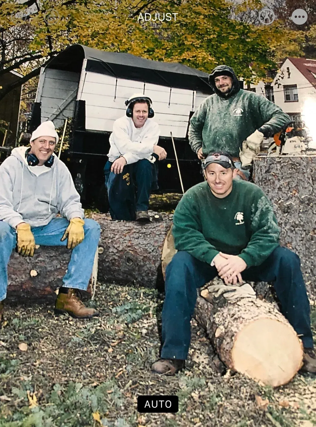 Four people posing outdoors beside a woodpile and trailer in autumn, wearing work clothes and smiling.