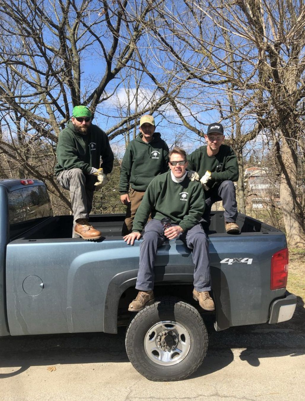 Four people in green hoodies posing on a gray pickup truck outdoors under bare trees.