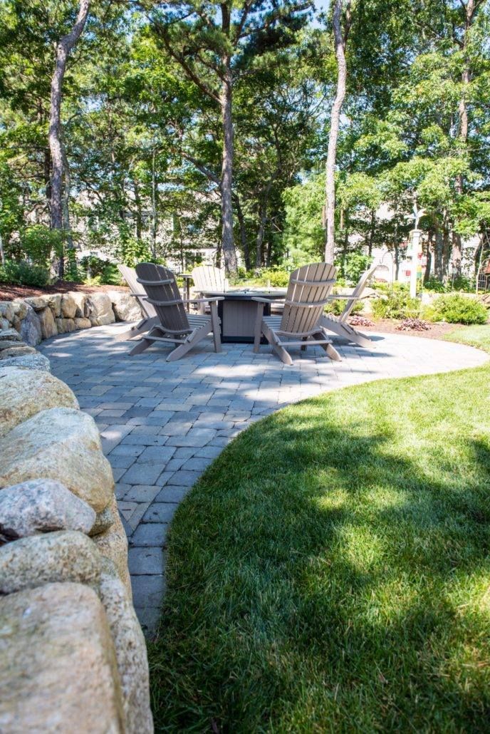 Patio with stone pavers, Adirondack chairs, and a table surrounded by green grass and trees.