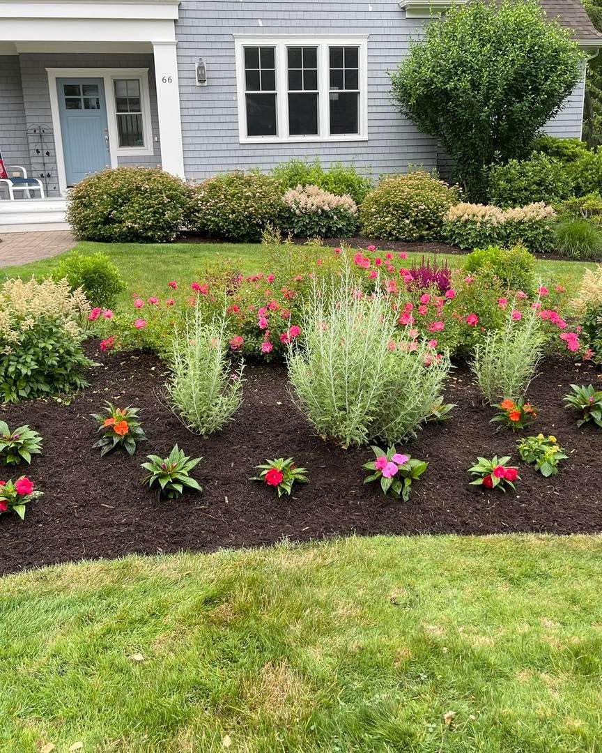 Flower garden in front of a gray house, with red, pink, and orange blooms and green bushes.