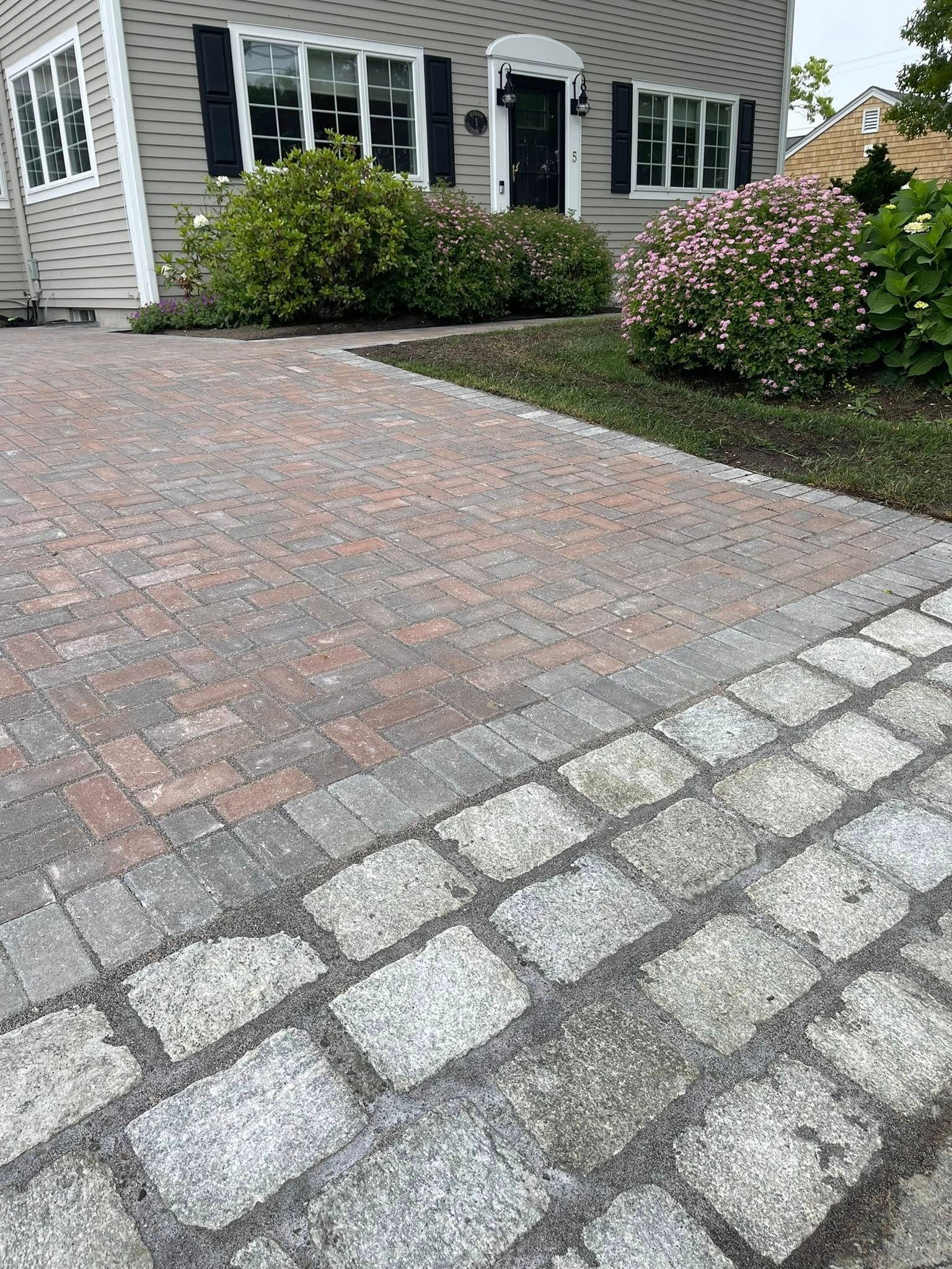 Brick driveway leading to a house with black shutters and a black door; bushes flank the path.