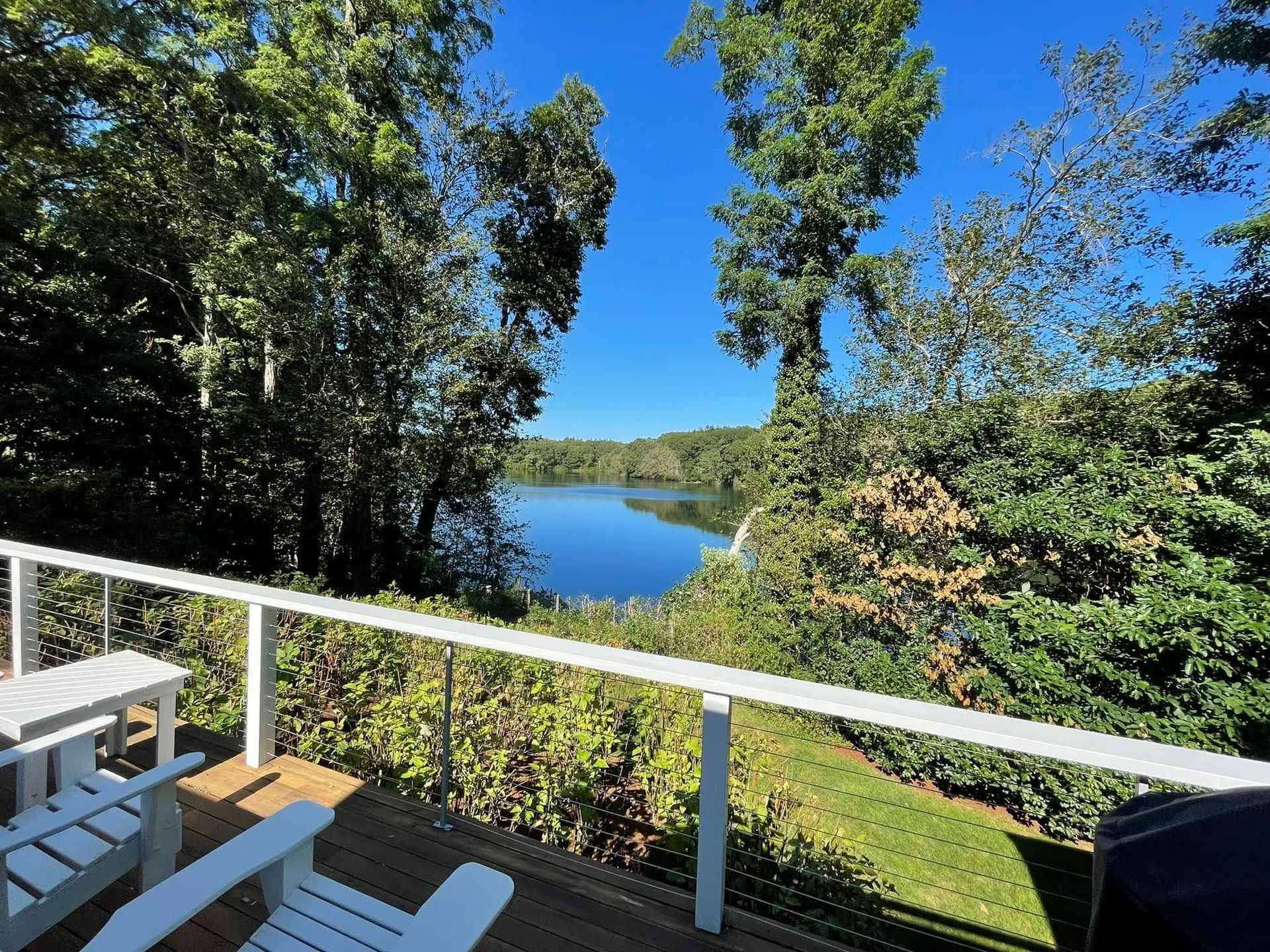 Deck overlooking a calm lake, framed by green trees and bright blue sky. Two white chairs on deck.