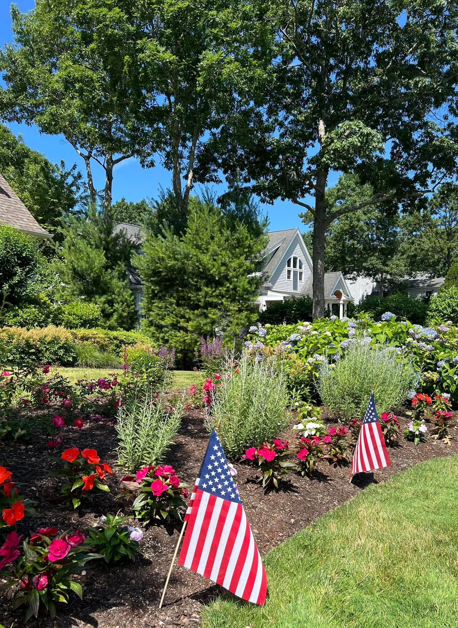 American flags in a colorful flower garden in front of a white house with trees under a bright blue sky.