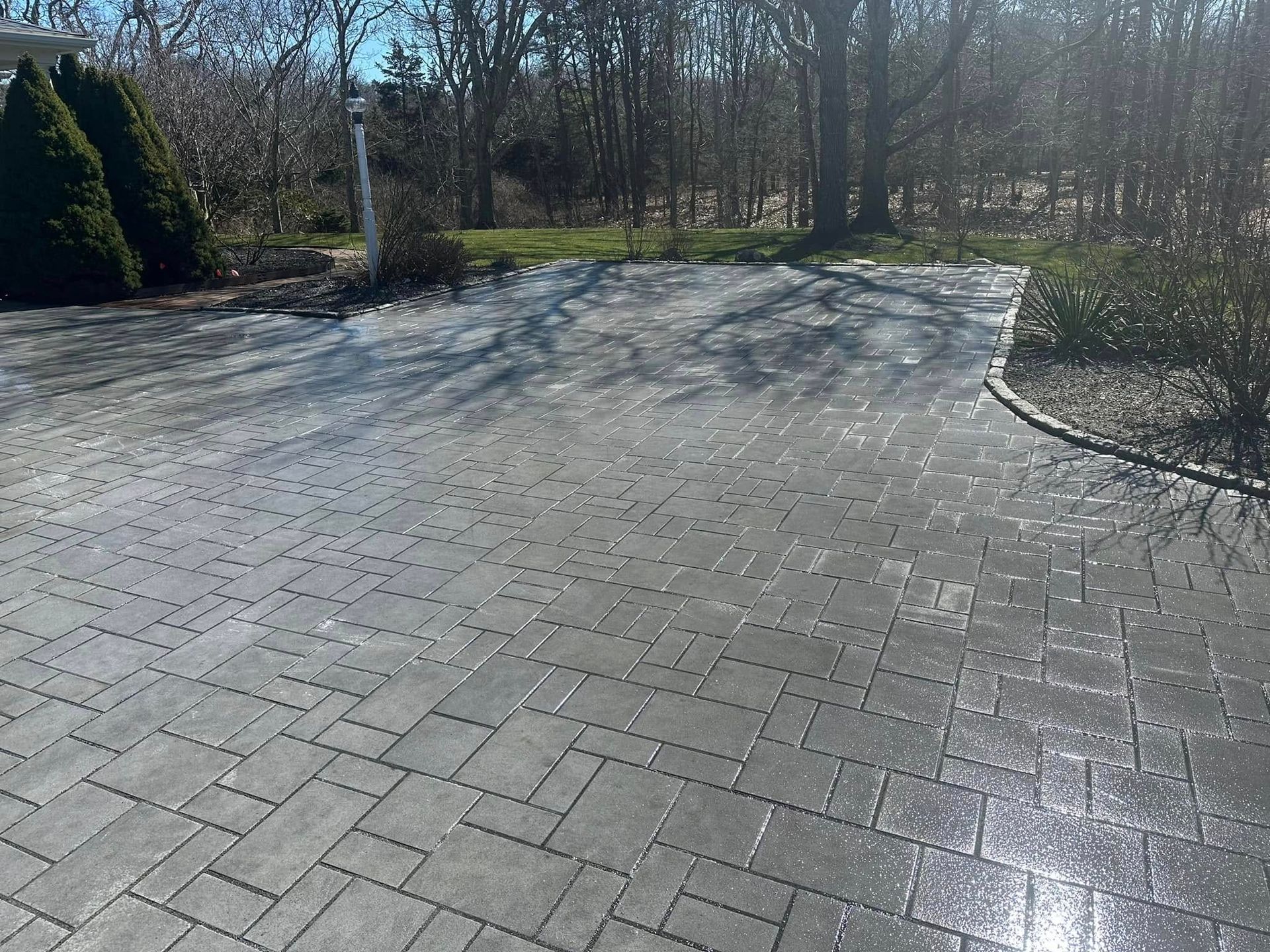 Gray brick paved driveway with glistening wet spots, trees in the background.