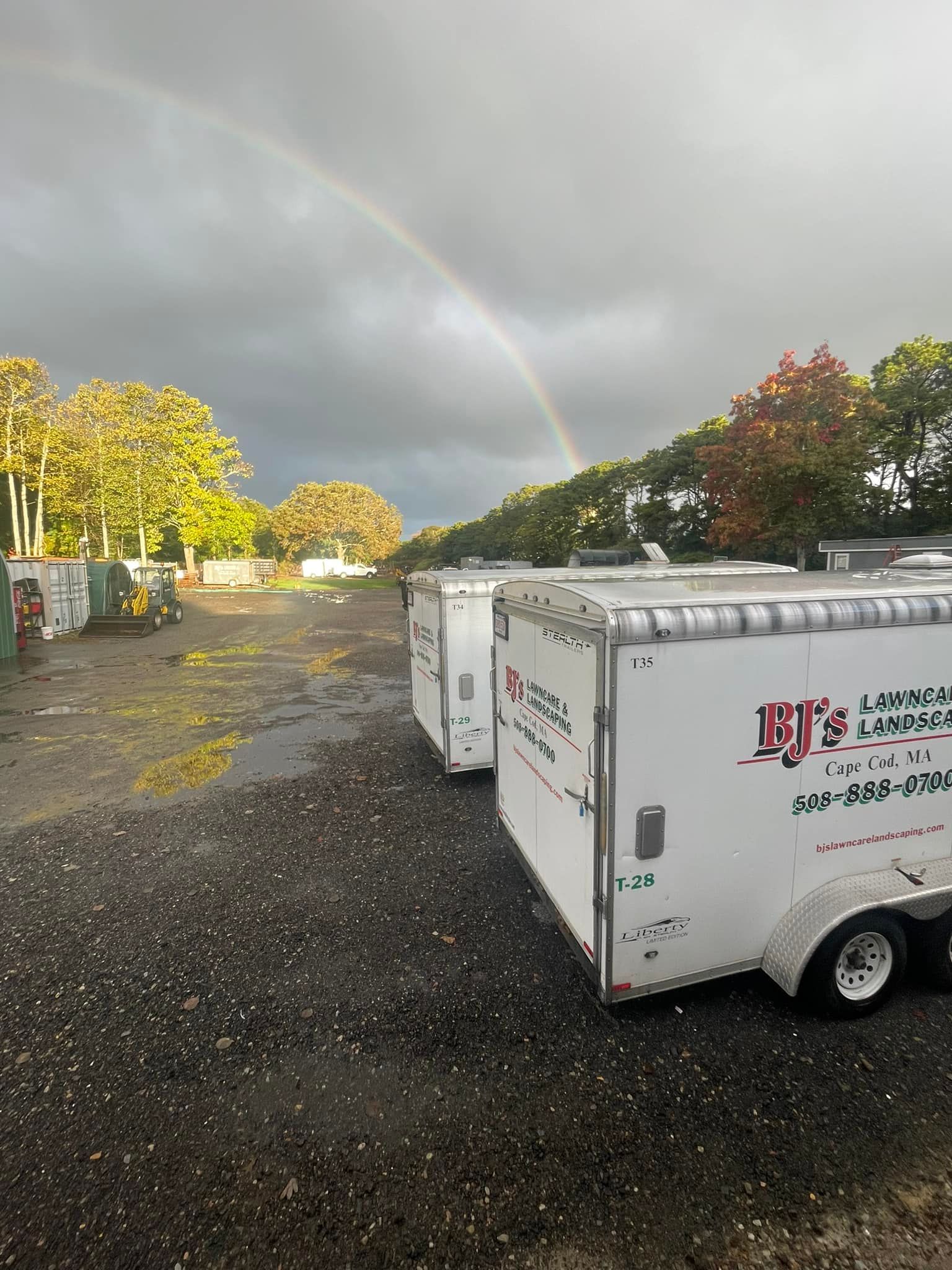 Two white trailers with red logos parked on a gravel lot under a rainbow.