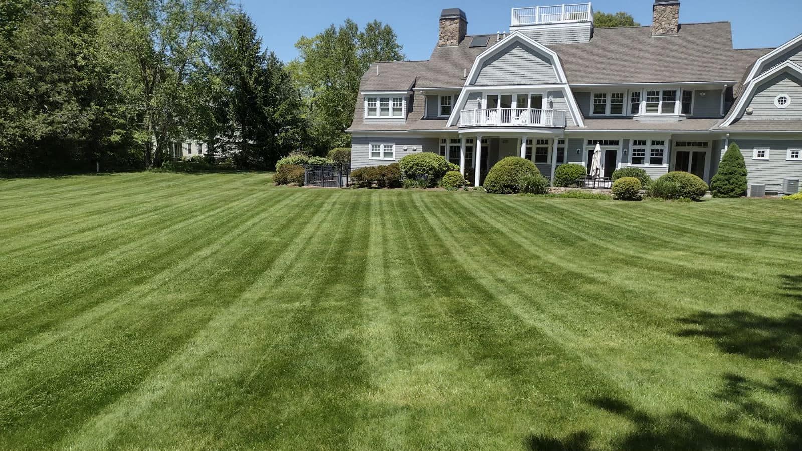 Green lawn with mowed stripes leading to a large gray house with a white balcony on a sunny day.