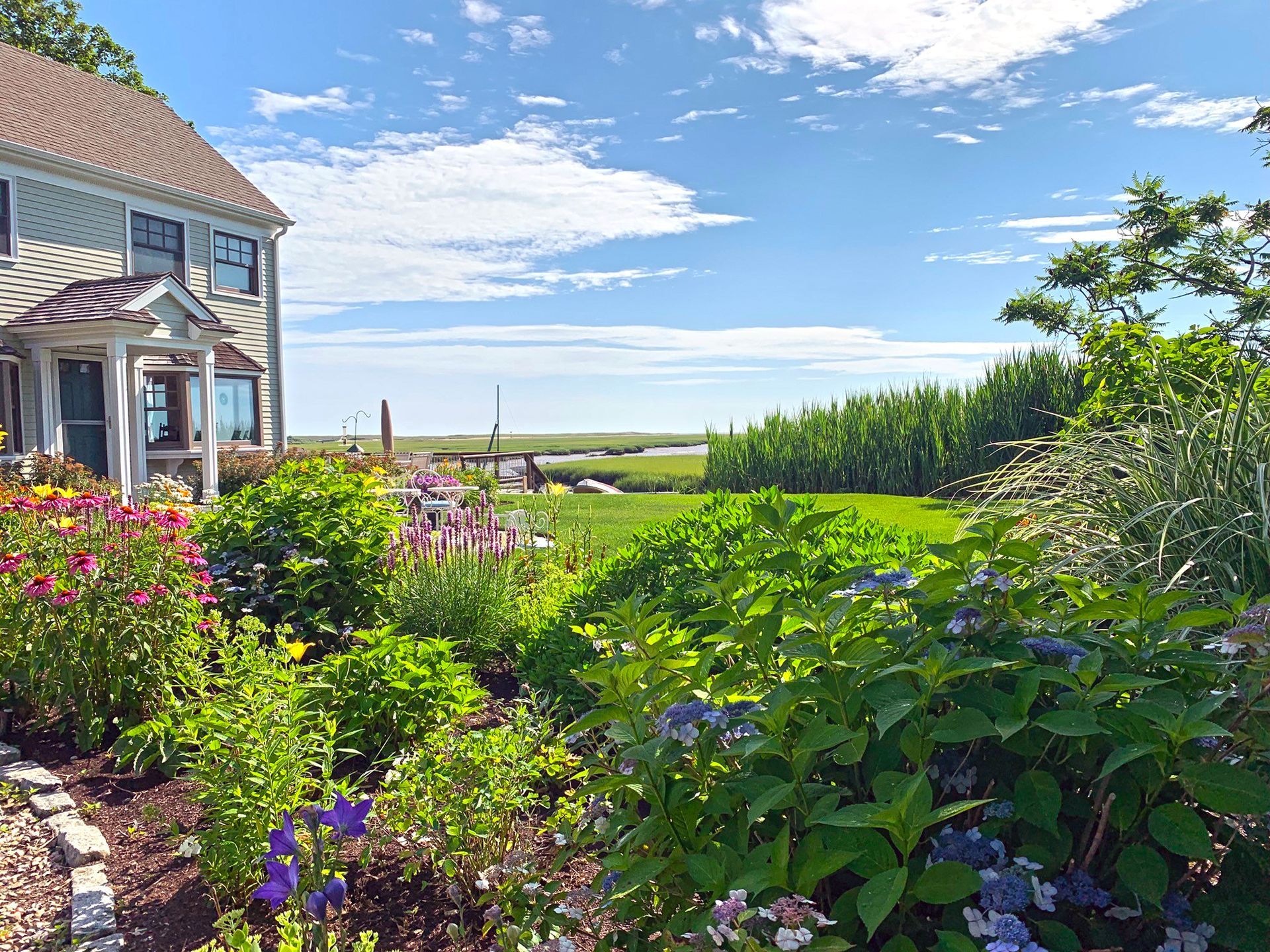 House overlooking a vibrant garden and waterfront, under a blue sky with clouds.