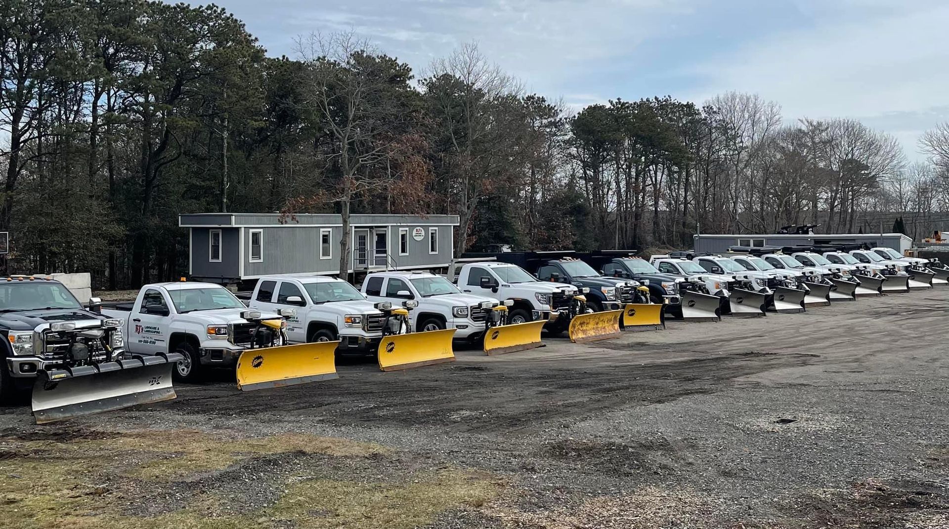 Line of white trucks with yellow snowplows parked on gravel in front of a small gray building.