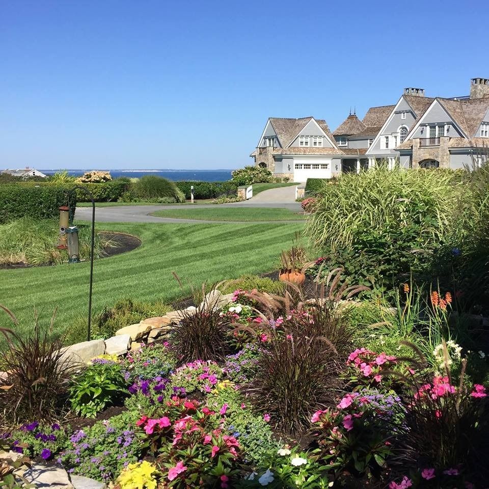 Lush garden in foreground with colorful flowers, green lawn with houses and ocean in background on a sunny day.