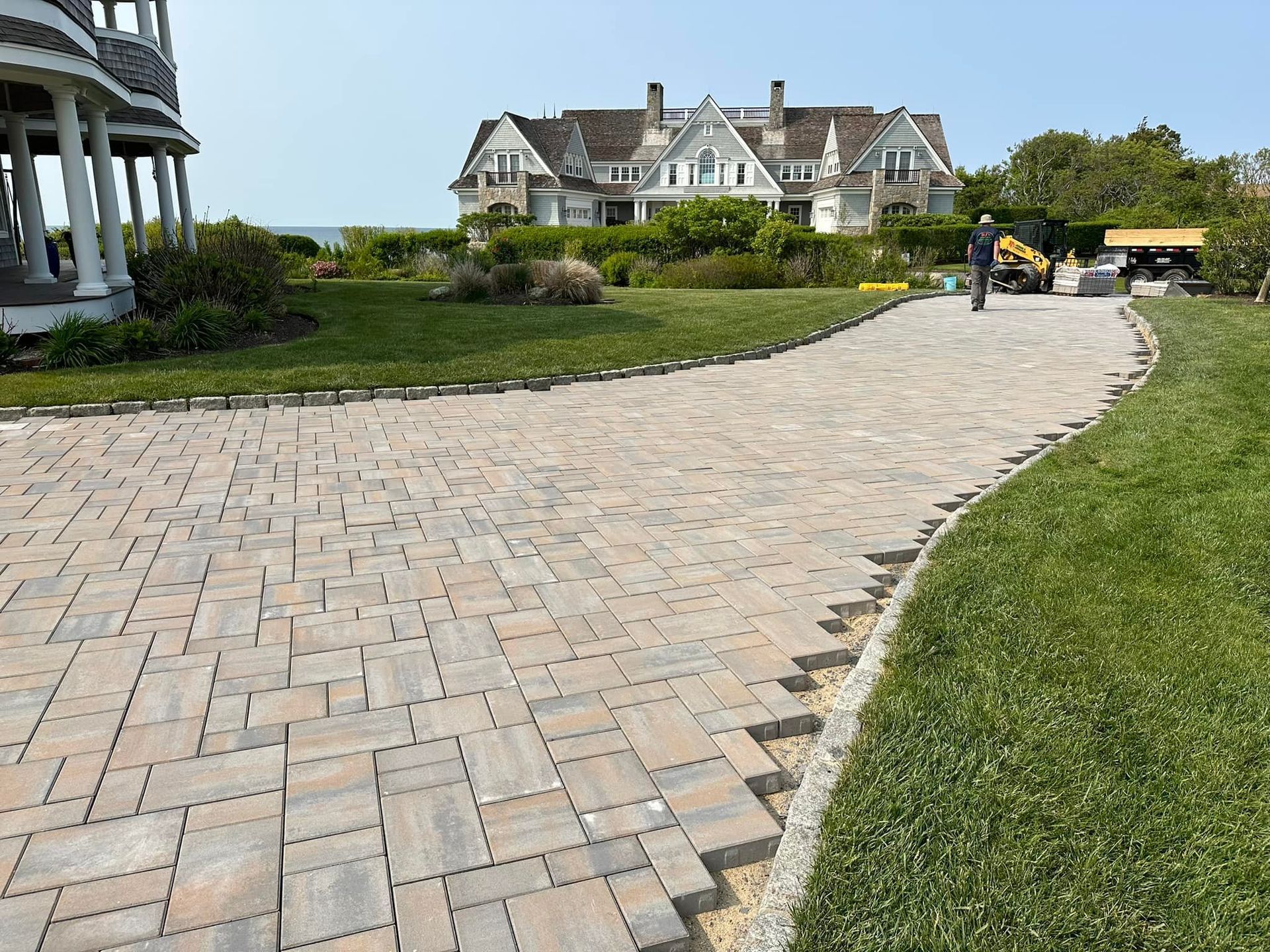 Brick paved driveway curving toward a large house, bordered by grass and stone edging.