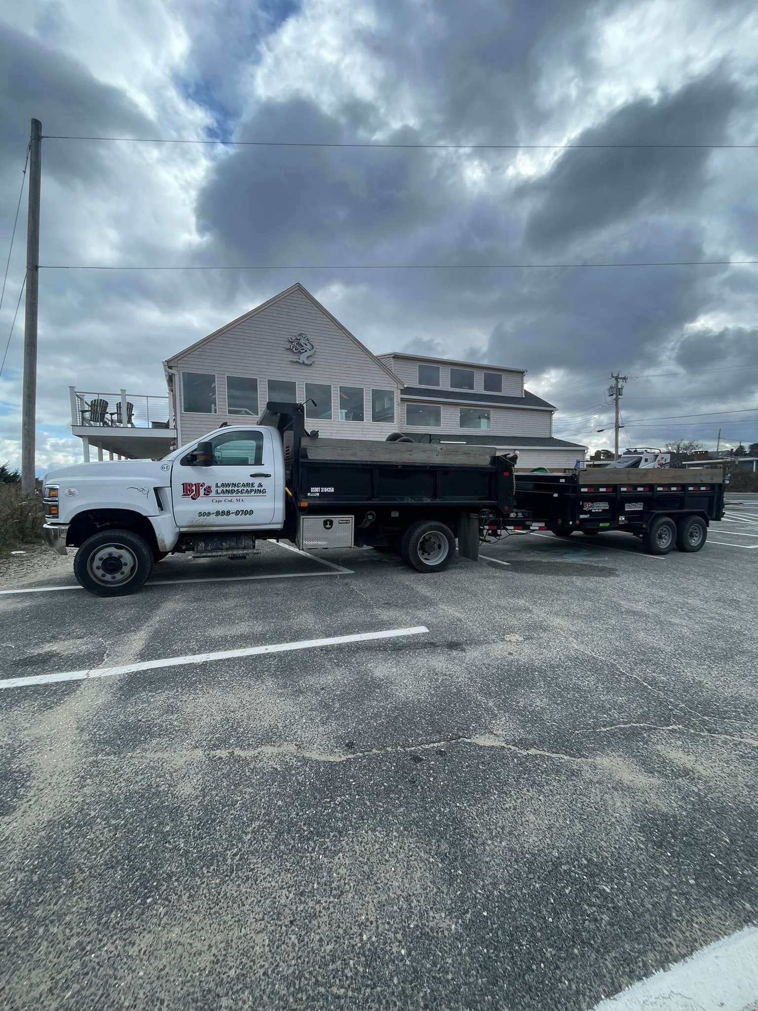 White dump truck with trailer parked outside a light-colored building under a cloudy sky.