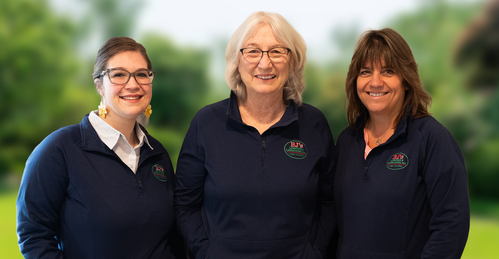 Three people wearing matching navy blue pullovers stand side-by-side against a soft, green outdoor background.