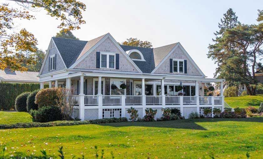 Gray-shingled house with white trim, covered porch, and green lawn on a sunny day.