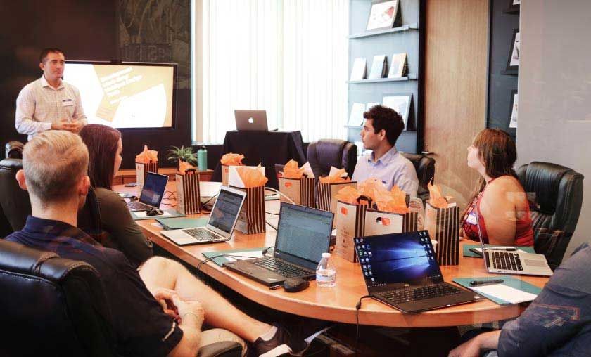 Man presenting at a conference table; laptops and gift bags are present, with people listening.