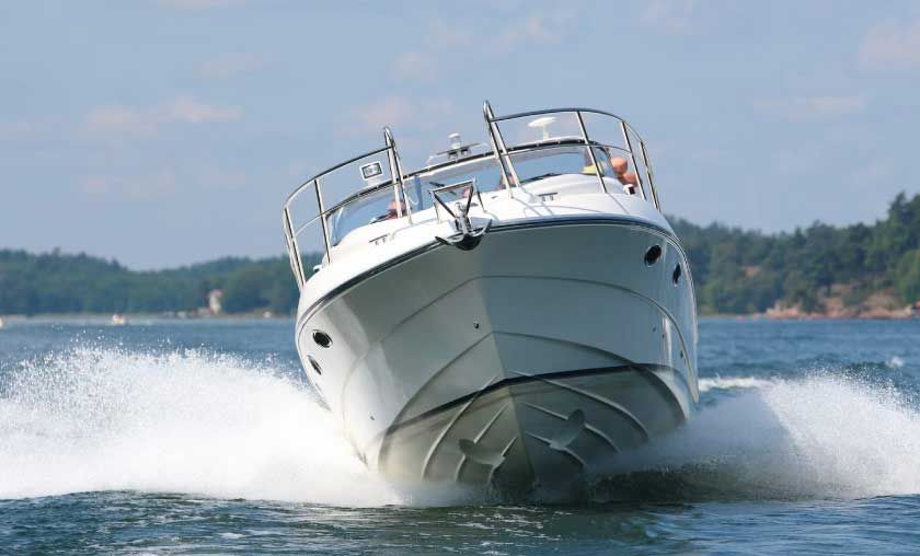 White motorboat speeding on water, creating a large wake. Blue sky and shoreline in the background.