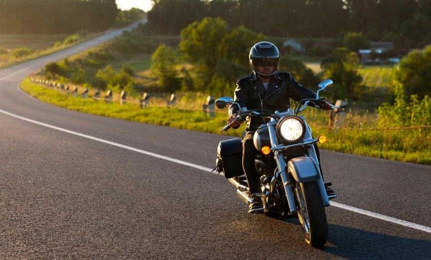 Motorcyclist on a winding road; wearing a black jacket and helmet, riding toward the viewer.