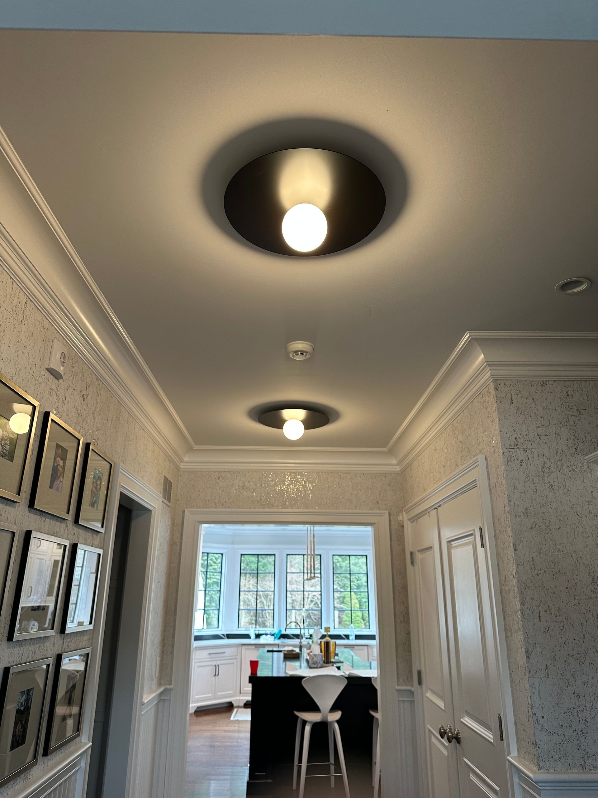 A hallway with three circular ceiling lights. White crown molding and speckled wallpaper line the walls. Kitchen visible at the end.