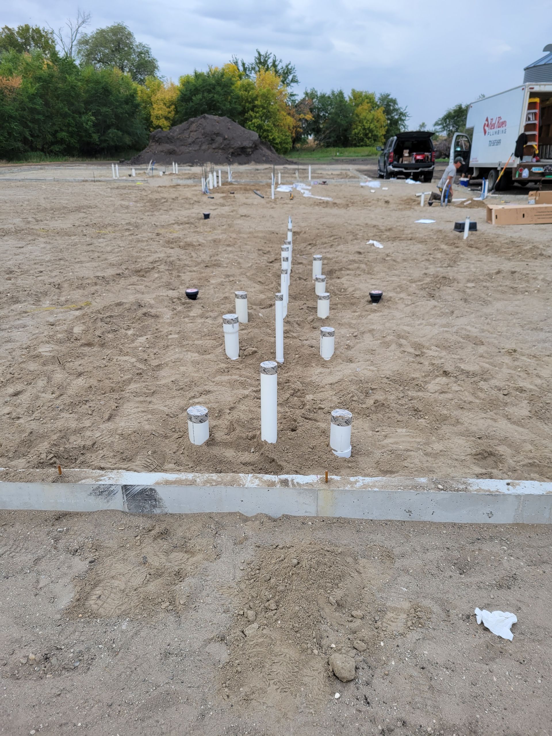 Concrete foundation footings laid out on a gravel lot with white PVC pipes and trucks in the background