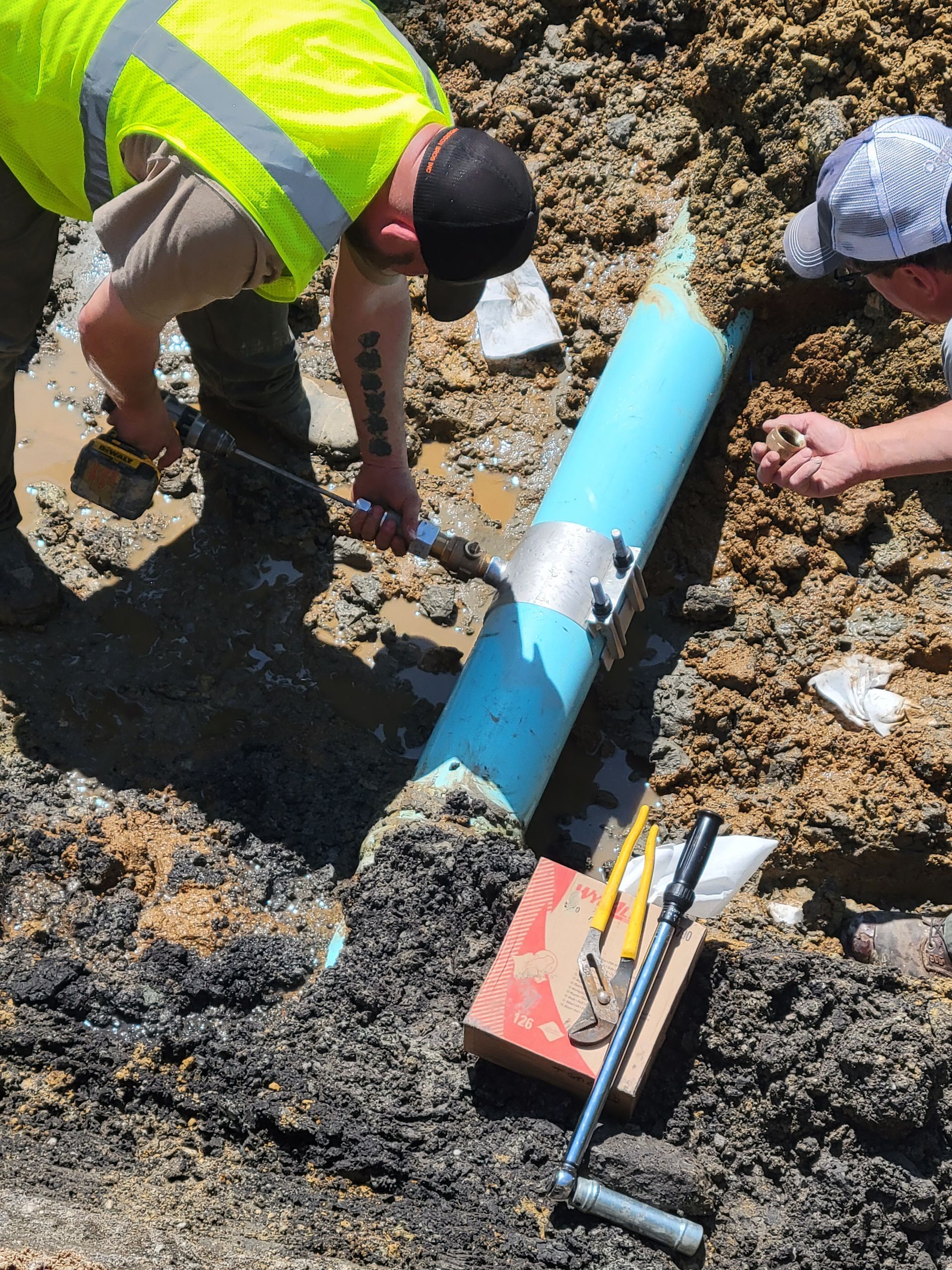 Workers repairing a blue pipe in a muddy trench with tools and a shovel nearby