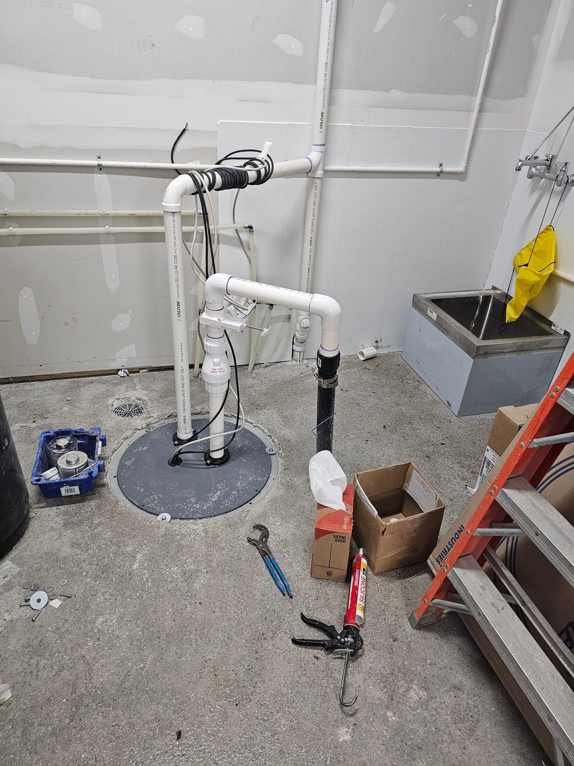 Plumbing work area with exposed white pipes, tools on the floor, and a ladder in a partially finished room