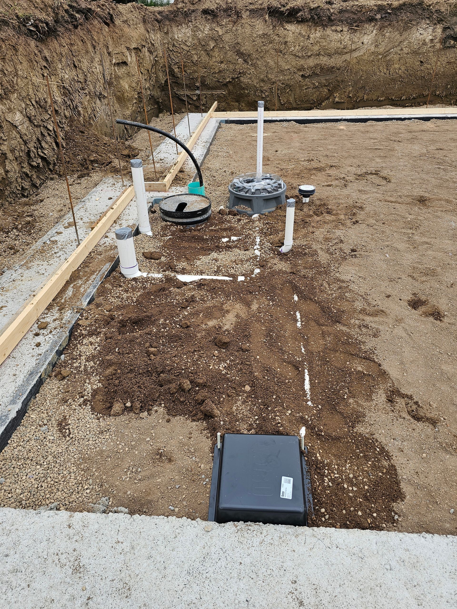 Outdoor utility site with white pipes, a round valve cover, and a black access cover on gravel.