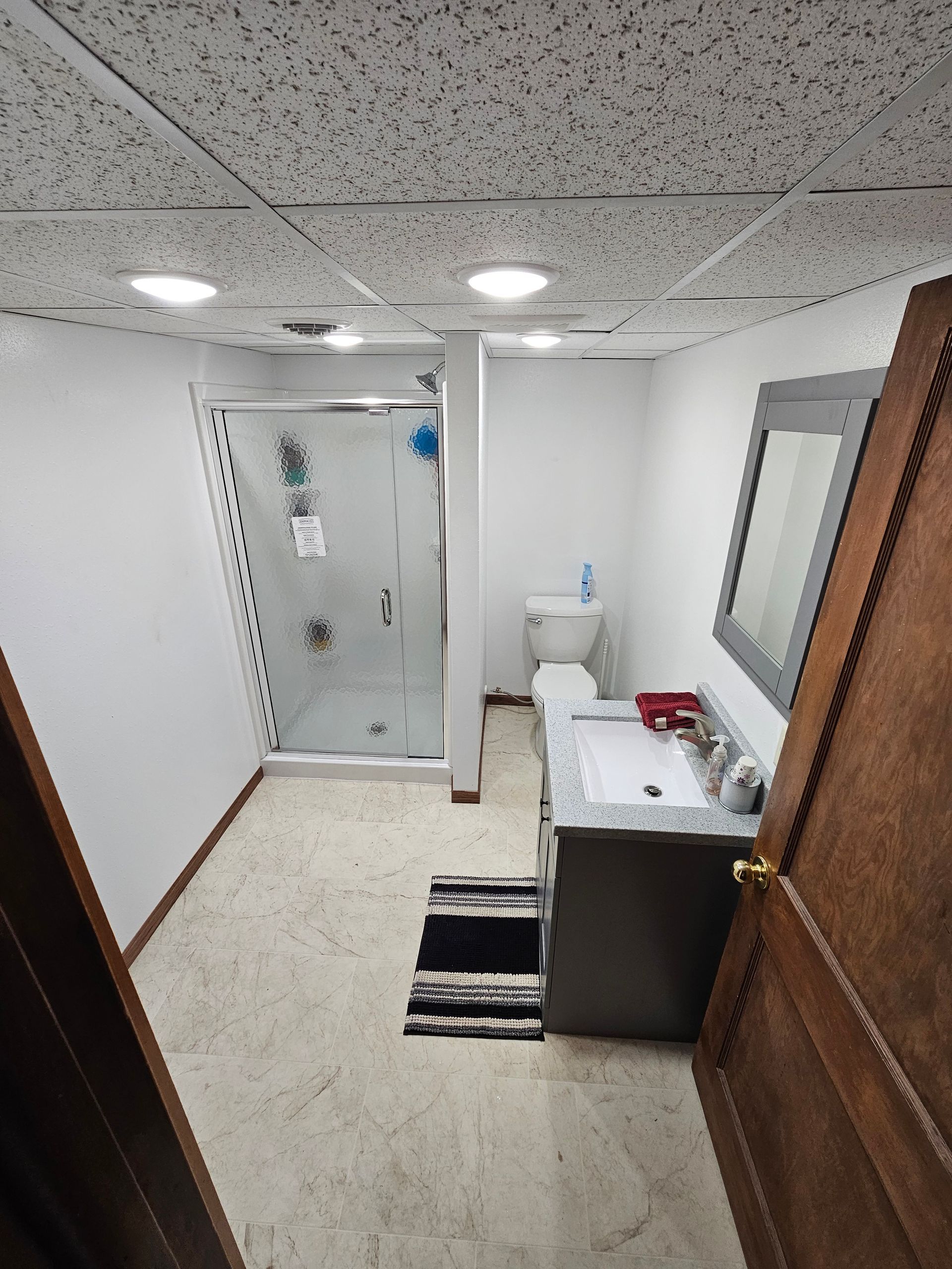 Small bathroom with shower, toilet, vanity, mirror, and striped rug viewed through an open wooden door