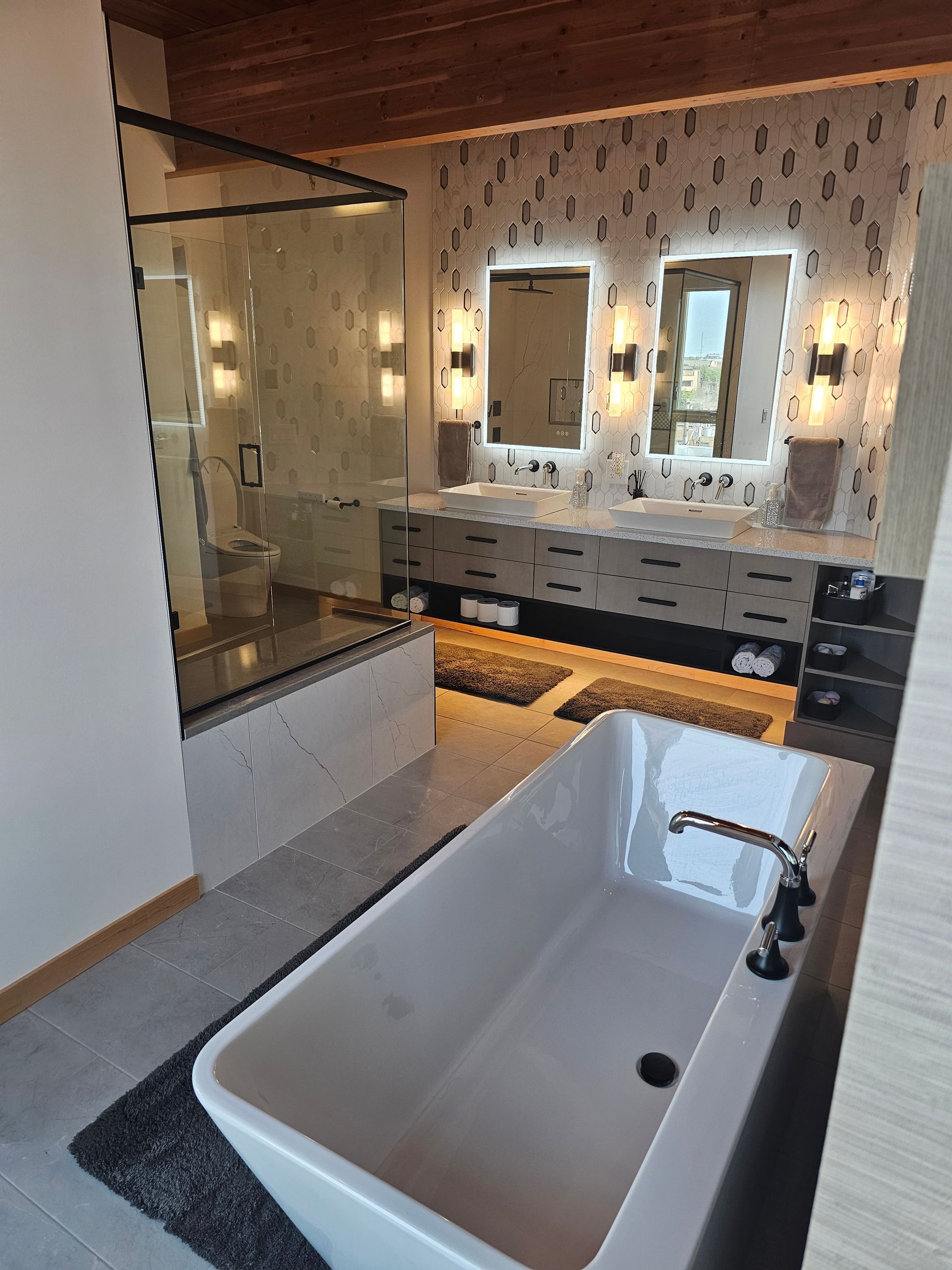 Modern bathroom with freestanding white tub, double vanity, mirrors, and warm wall sconces.