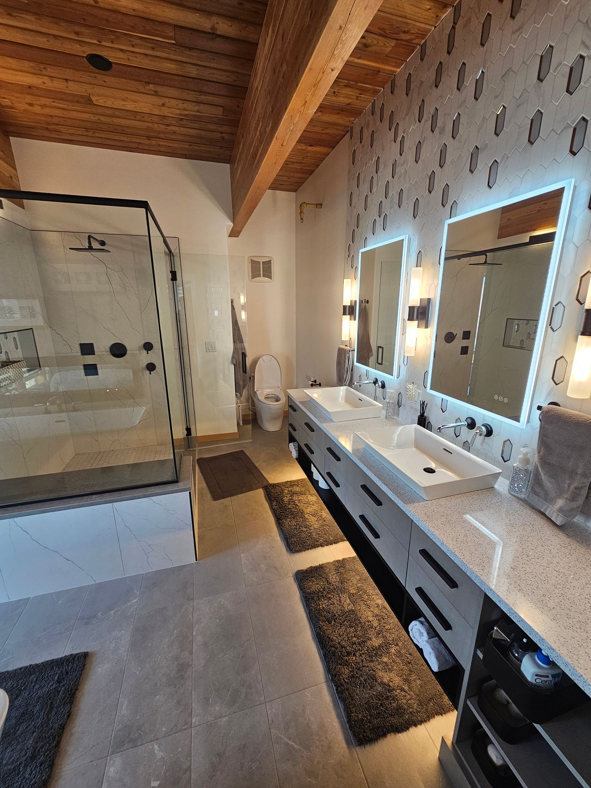 Spacious bathroom with double vanity, illuminated mirrors, glass shower, and wood-beamed ceiling.