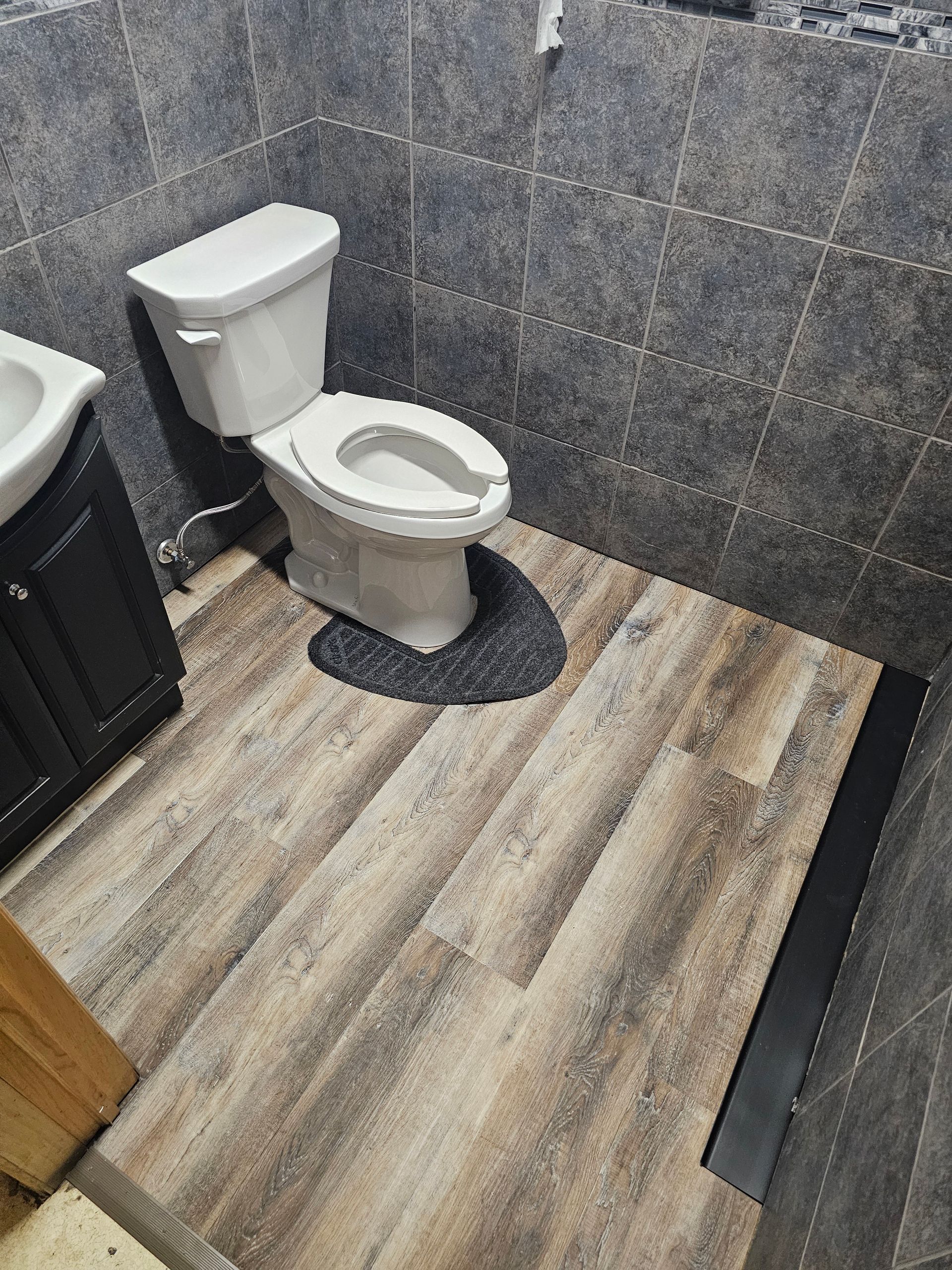 Small bathroom with white toilet, dark gray tile walls, and wood-look vinyl flooring.