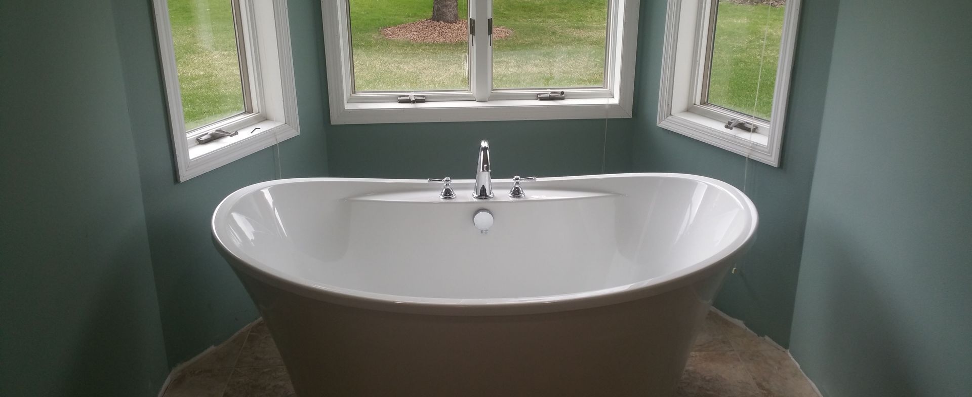 White bathtub beneath windows in a green bathroom, with a chrome faucet centered on the rim