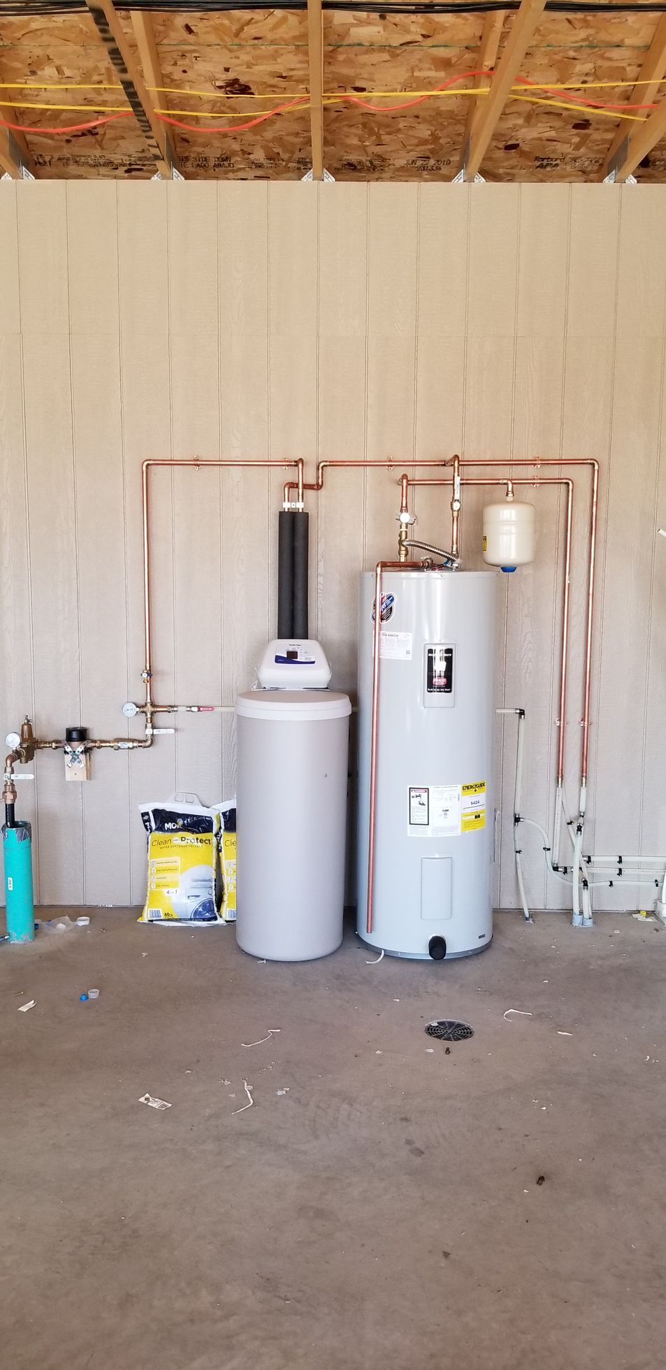 Water heater and softener setup in a garage with exposed ceiling beams and utility pipes.