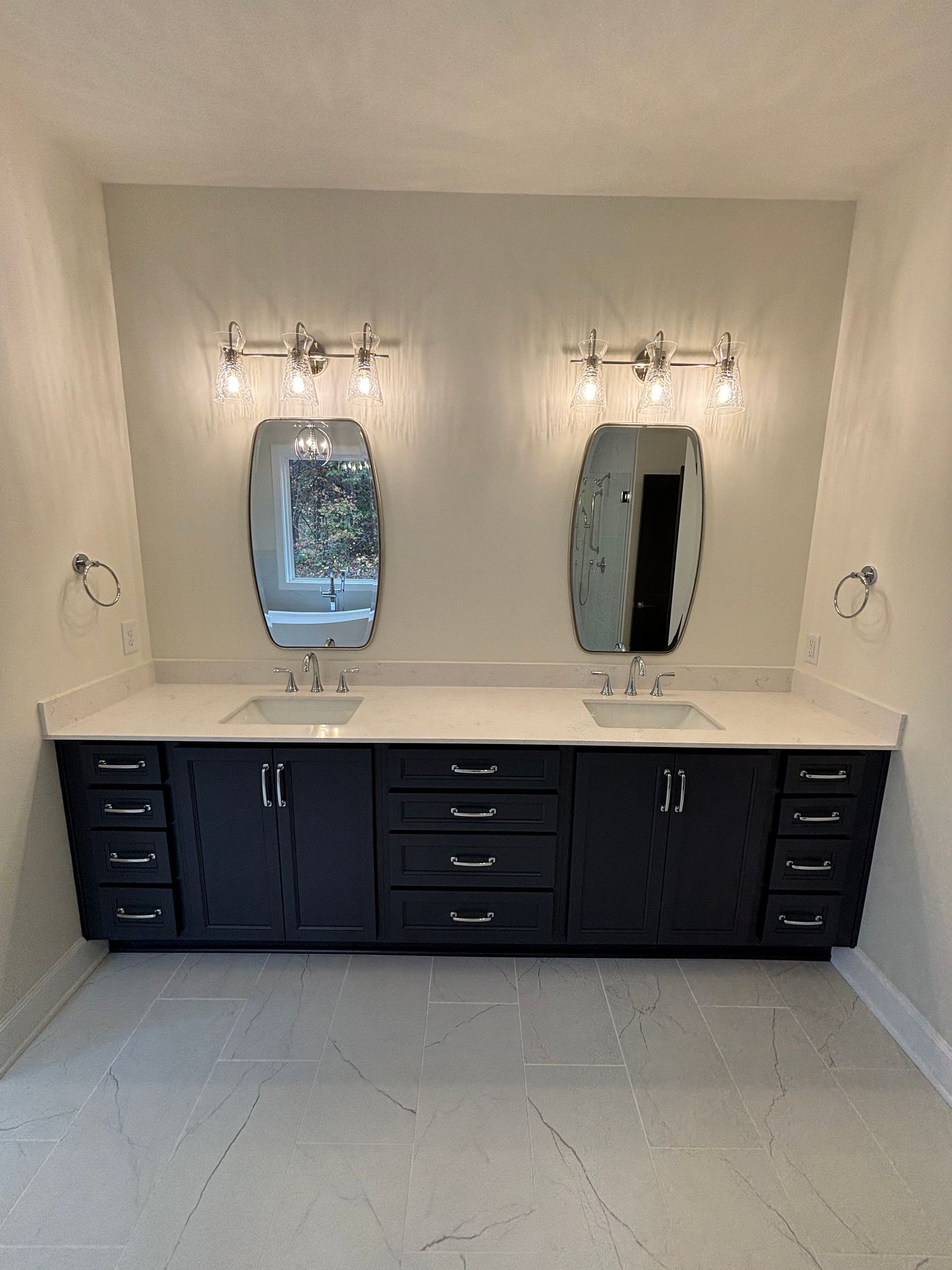 A modern bathroom with a double sink vanity. Black cabinets, white countertop, oval mirrors, and sconce lighting.