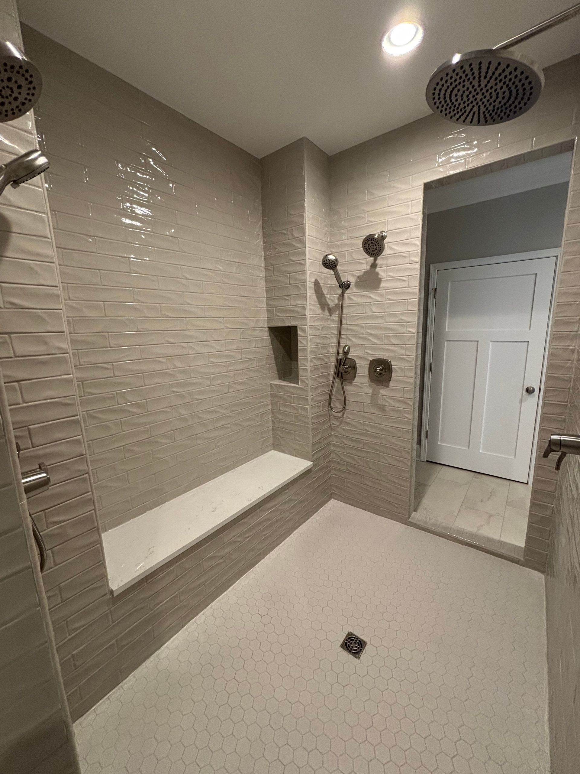 Bathroom shower with light beige textured tile walls, white hexagonal floor, and a bench.
