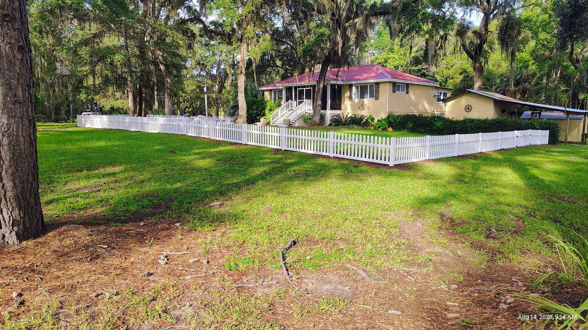 A small, yellow house with a red roof sits behind a white picket fence in a wooded area.