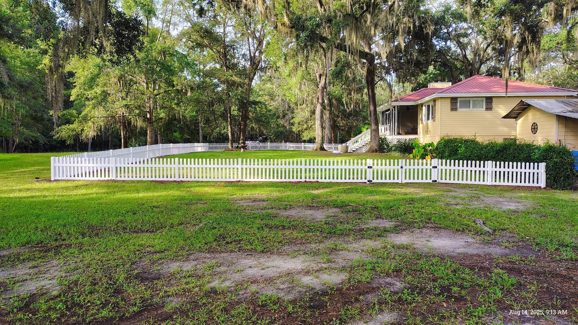 White picket fence surrounds a grassy yard, with a yellow house and trees in the background.