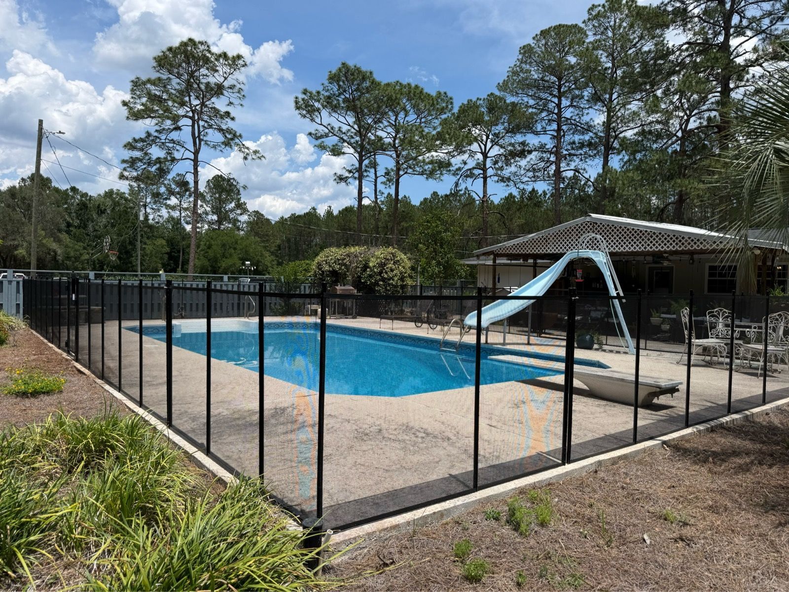 Fenced backyard pool with a blue slide and screened patio under tall pine trees.